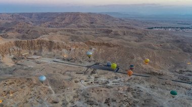 Parlak balonlar Luxor 'daki arkeolojik bölgede uçuyor. Aşağıda kum tepeleri, antik tapınaklar, devam eden kazılar var. Şafak vakti gökyüzünün arka planına karşı pitoresk bir dağ sırası. Mısır