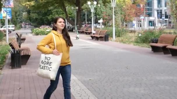 En brunett kvinna i en orange luvtröja går med en tygpåse som säger Ingen mer plast i staden — Stockvideo