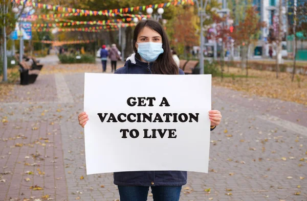 Naisella lääketieteellisessä naamiossa on juliste, jossa sanotaan: "Hanki rokotus elääksesi". — kuvapankkivalokuva
