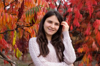 25 yaşında esmer bir kadın uzun saçlarını düzeltir ve sonbaharda kırmızı yaprakların arka planına gülümser.