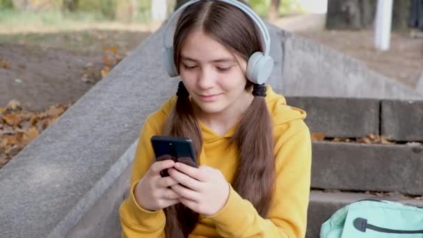 En tonårstjej i hörlurar skriver på en smartphone och ler, sms:ar med en vän. — Stockvideo