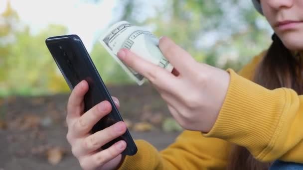 De tiener heeft dollarbiljetten en kijkt naar het scherm van de smartphone. — Stockvideo