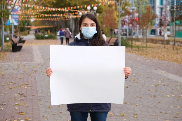 Una persona con una máscara médica protectora sostiene una hoja blanca vacía de papel sin texto. — Foto de Stock