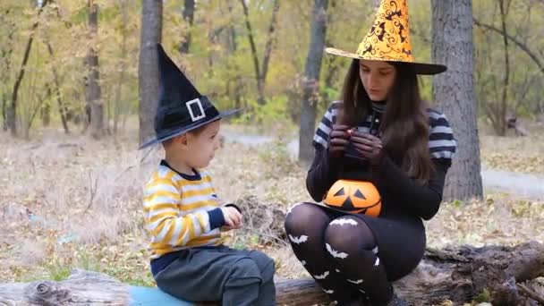 Matka a syn v kostýmech Halloween jíst oranžové bonbóny v podzimním lese. — Stock video