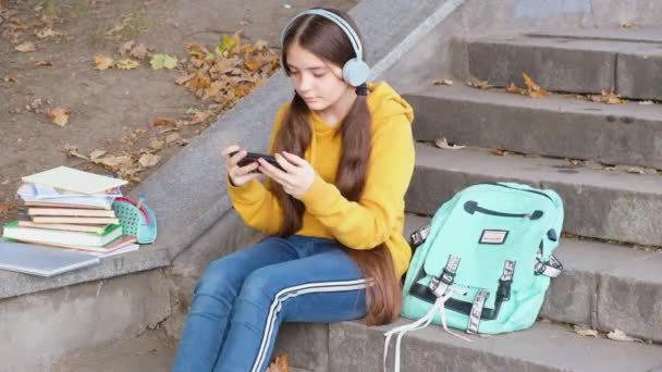 Una adolescente con auriculares mira seriamente en la pantalla del teléfono inteligente, aprendizaje en línea. — Vídeos de Stock