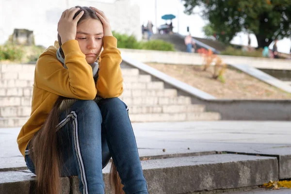 A sad teenage girl sits on the stairs and covers her head with her hands — Stock Photo, Image