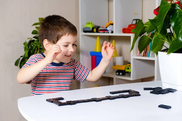 Um menino feliz de 4 anos joga um jogo de tabuleiro de dominós, se diverte e gosta do jogo — Fotografia de Stock