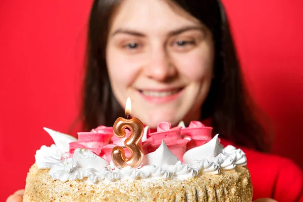 Una mujer feliz sostiene un pastel con una vela en la forma del número tres sobre un fondo rojo. —  Fotos de Stock