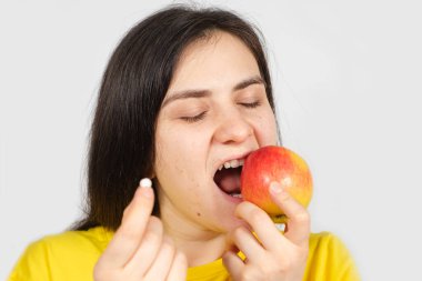 Bir kadın elinde multivitamin hapı tutarken kırmızı bir elmayı ısırıyor.