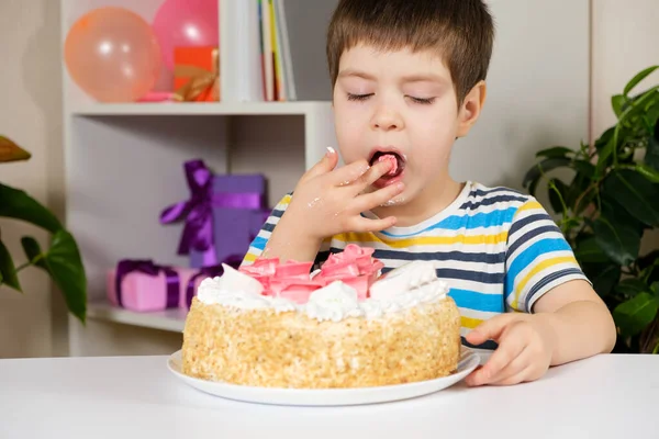 Un niño feliz come un pastel de cumpleaños, come con sus manos y se lame los dedos. — Foto de Stock