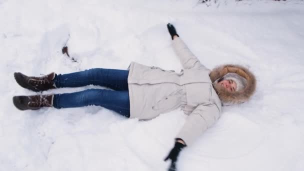 Una mujer con una chaqueta blanca yace en la nieve, levanta las manos hacia arriba y hacia abajo, como si agitara sus alas y hiciera un ángel — Vídeos de Stock