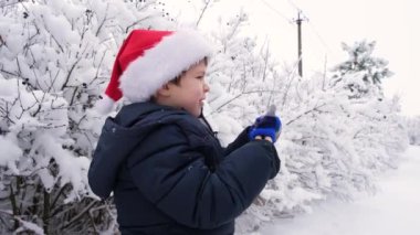 4 yaşında, Noel Baba şapkalı yakışıklı bir çocuk karlı bir ormanda eldivenleri takıyor.