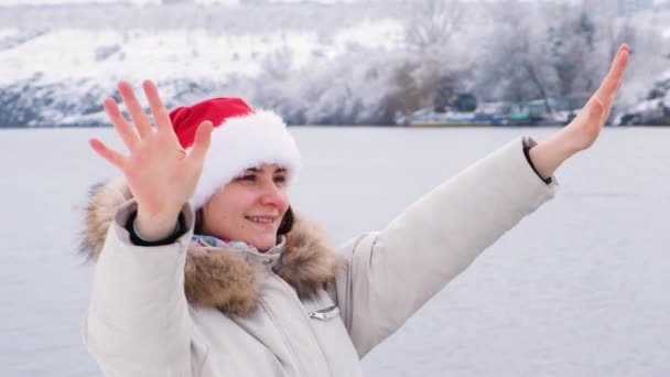 Un video retrato de una hermosa mujer en sombrero de Santas, que camina en la naturaleza a lo largo del río, levanta las manos y sonríe — Vídeo de stock