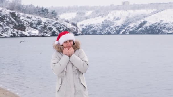 Una mujer en el sombrero de Santas camina a lo largo del río en la naturaleza en invierno, el estado de ánimo de Navidad, cámara lenta. — Vídeos de Stock