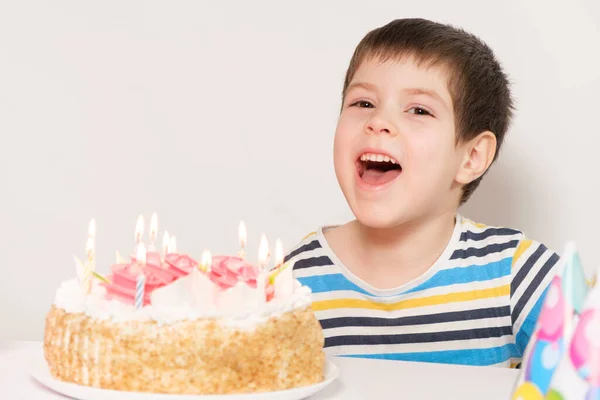 Un niño se ríe, celebra su cumpleaños, se sienta frente a un pastel blanco con velas. — Foto de Stock