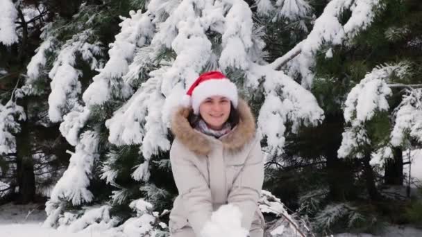 Una mujer en el sombrero de Santas vomita nieve en un bosque de pinos de invierno, el estado de ánimo de Navidad. — Vídeo de stock