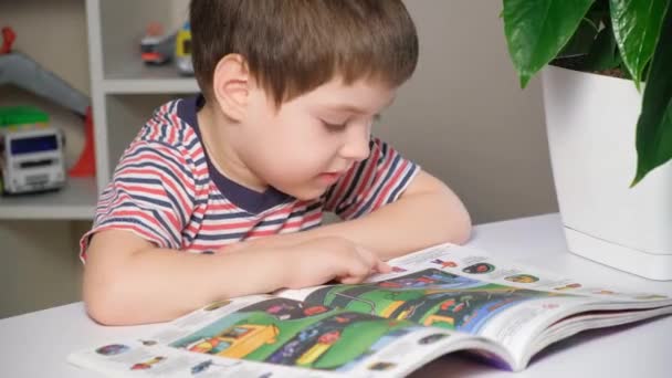 Um menino pré-escolar aprende a ler, olha para fotos em um livro e fala. — Vídeo de Stock