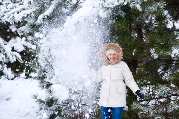 Una mujer con una chaqueta blanca en un bosque de pinos de invierno salta y se divierte bajo ramas cubiertas de nieve bajo nieve — Foto de Stock