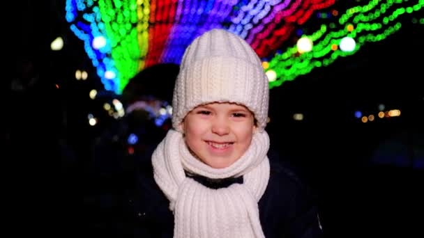 A Christmas kid in a white hat and scarf smiles as he stands under garlands in the winter and walks forward looking into the camera. — Stockvideo