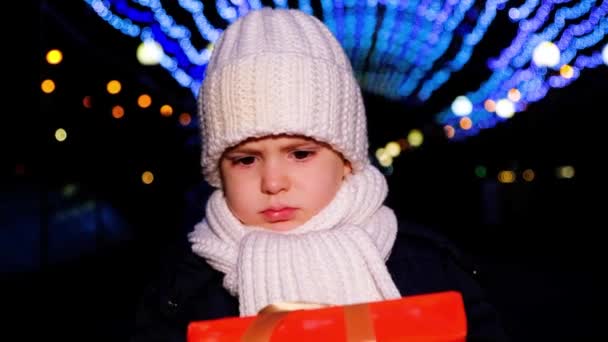 Een kleine jongen is verdrietig voor Kerstmis, met een geschenk tegen de achtergrond van een glanzende bloemenslinger — Stockvideo