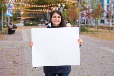 25 yaşında esmer bir kadın gülümsüyor ve elinde yazı ve yazı için boş beyaz bir çarşaf tutuyor.