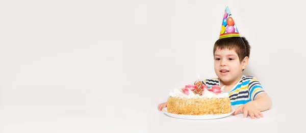 Un enfant dans une casquette festive regarde un gâteau avec une bougie sous la forme du numéro trois. — Photo