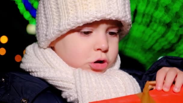A little boy in a white hat and scarf stands under garlands on Christmas Day and holds a box of gifts. — Vídeo de Stock