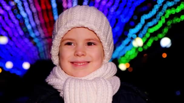 A handsome child in a white hat smiles standing under Christmas garlands in the evening — Vídeo de Stock