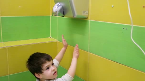 Seorang anak laki-laki berusia 3-4 tahun mengeringkan tangannya dengan pengering otomatis listrik — Stok Video
