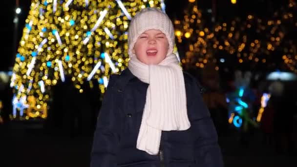 Een prachtig kleuter glimlacht terwijl hij in de camera kijkt en 's nachts bij een kerstboom staat. — Stockvideo
