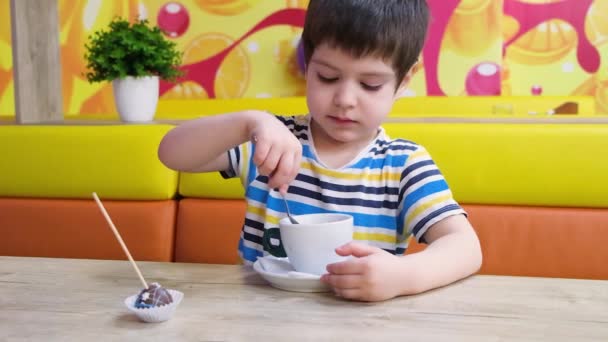 Een 4-jarige jongen in een gestreept T-shirt drinkt cacao van een lepel in een café.. — Stockvideo