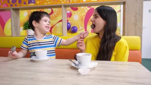 Mãe e filho comem na sala de jogos, bebem cacau e comem chocolates, se alimentam. — Vídeo de Stock