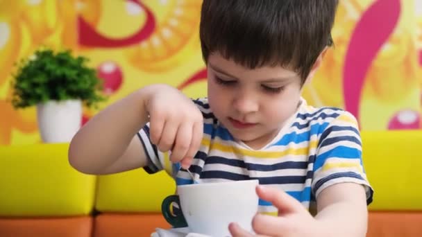 En 4-årig pojke i randig t-shirt dricker kakao från en sked på ett kafé. — Stockvideo