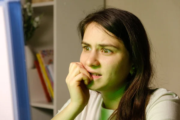 A woman carefully looks at the TV or computer screen, bites her nails, watches a movie or TV series. — Stock Photo, Image