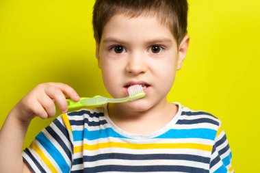 Bir çocuk elinde pembe diş macunuyla bir diş fırçası tutuyor, sarı arka plandaki kameraya bakıyor.