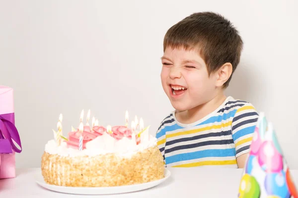 Un niño se ríe, celebra su cumpleaños, se sienta frente a un pastel blanco con velas. — Foto de Stock