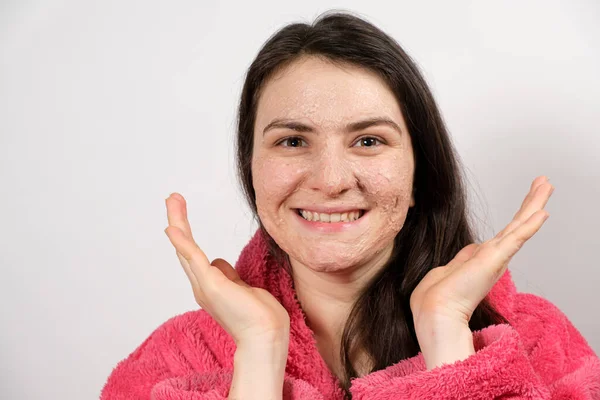 Una joven morena con un albornoz rosa hace tratamientos faciales limpiadores: una máscara o un exfoliante para limpiar y nutrir la piel. —  Fotos de Stock