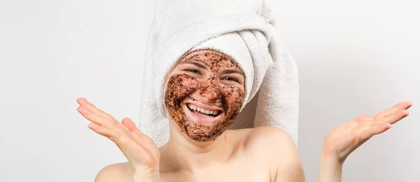 Μια νεαρή γυναίκα με μια μάσκα καφέ στο πρόσωπό της και μια πετσέτα μπάνιου στο κεφάλι της χαμογελά και κοιτάζει στην κάμερα σε ένα λευκό φόντο, ένα μέρος για κείμενο. — Φωτογραφία Αρχείου