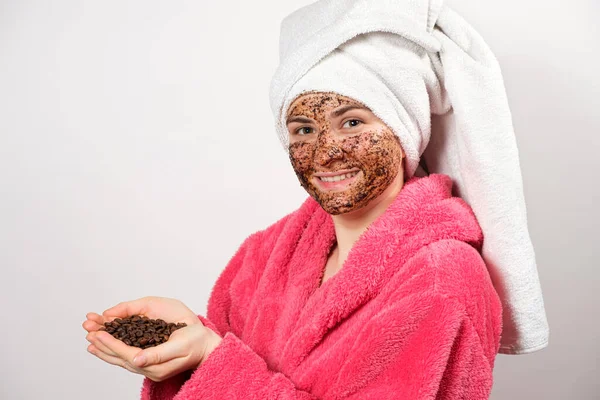 Una mujer con un albornoz rosa con una máscara de café en la cara sostiene granos de café en sus manos —  Fotos de Stock
