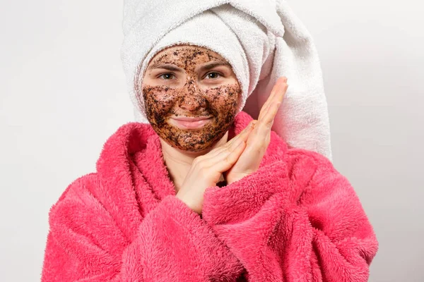 Egy gyönyörű nő, kávéskannával az arcán, fürdőköpenyben, törülközővel a fején, fehér alapon, gyógyfürdős bőrápolási eljárásokat tesz lehetővé. — Stock Fotó