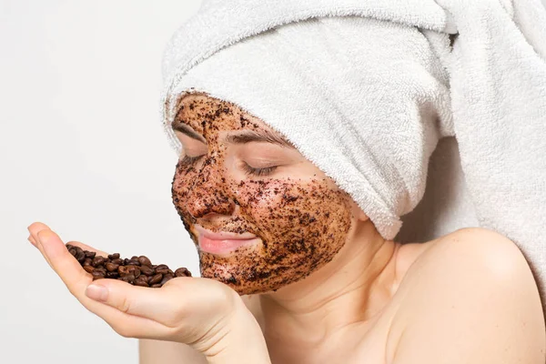 Une femme avec un masque de café ou un gommage sur son visage tient une poignée de grains de café dans sa main — Photo