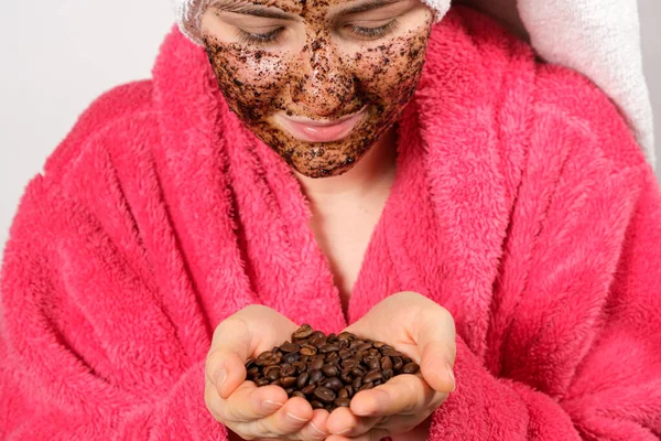 Una mujer con un albornoz rosa con una máscara de café en la cara sostiene granos de café en sus manos —  Fotos de Stock