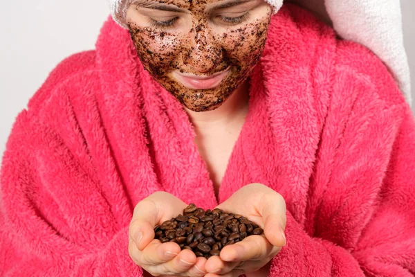 Een vrouw in een roze badjas met een koffiemasker op haar gezicht houdt koffiebonen in haar handen — Stockfoto