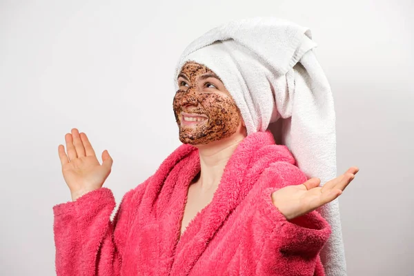 Een mooie vrouw met een koffie scrub op haar gezicht in een badjas met een handdoek op haar hoofd op een witte achtergrond maakt spa huidverzorging procedures — Stockfoto