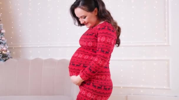 Eine schöne schwangere Frau in einem roten Winterkleid streicht über ihren Bauch vor weißem Hintergrund, ein gemütlicher Schimmer von Girlanden. — Stockvideo