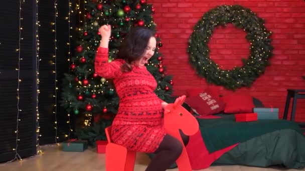 Een gelukkige zwangere vrouw rijdt op een rood houten speelgoedpaard en heeft plezier voor te bereiden op Kerstmis. — Stockvideo