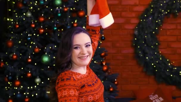 Video porträtt av en vacker brunett flicka i en röd klänning med en jultomte hatt på bakgrunden av en julgran. — Stockvideo
