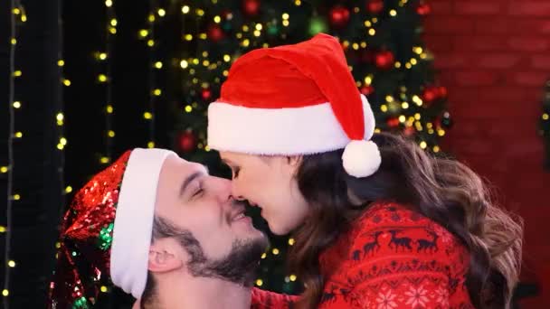 Ein glücklicher Kerl und ein Mädchen mit roten Nikolausmützen küssen sich und haben Spaß vor dem Hintergrund der schimmernden Girlanden des Weihnachtsbaums. — Stockvideo