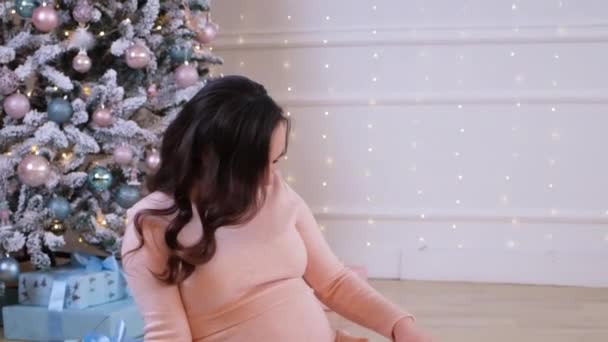 幸せな妊婦はピンクのドレスでクリスマスツリーの近くに座って、彼女の腹をストロークし、妊娠中の女性のためのクリスマスの販売. — ストック動画