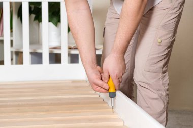 Mobilya derleyicisi yatağı monte eder, tahtadan levhaların vidalarını çevirir, elleri yakın çekim tornavidayla çevirir.